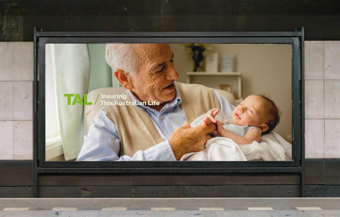 An image of a billboard at a train station, featuring an older man holding a baby. The words "TAL - Insuring This Australian Life" are superimposed. 