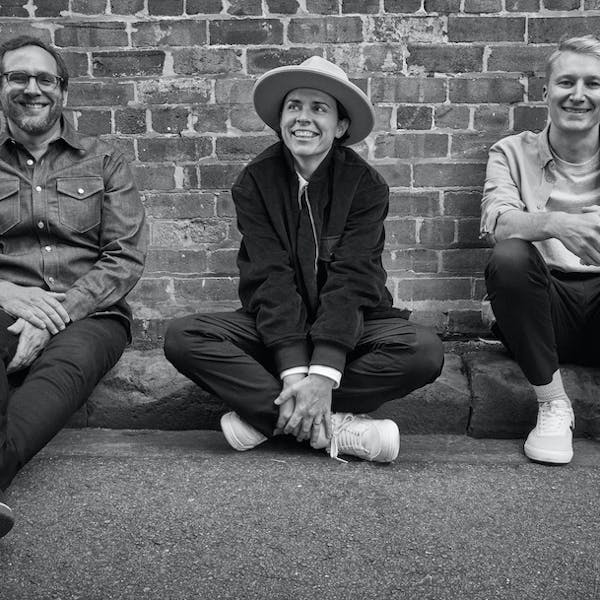 A black and white image of three people with cheerful expressions sitting against a brick wall.
