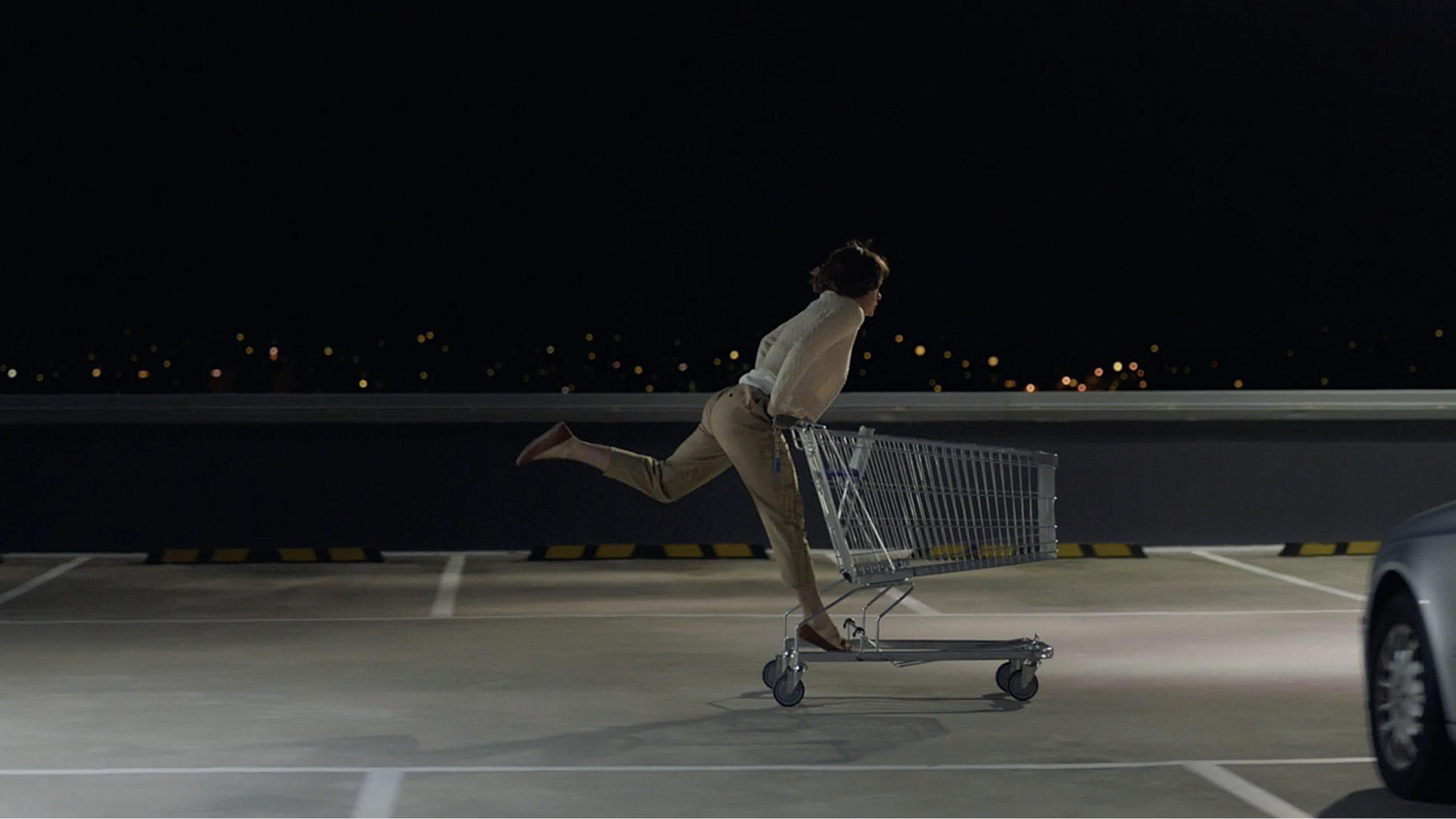 A still from a television commercial. A woman is standing on the back of a shopping trolley, rolling through a deserted carpark at night. 