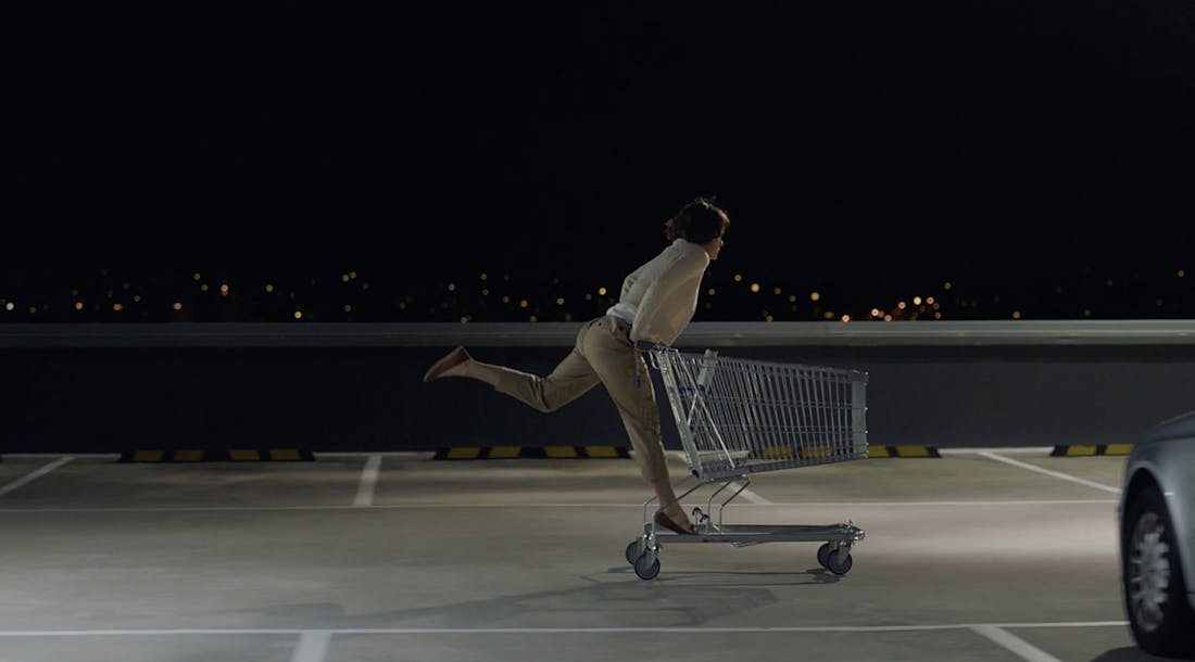 A still from a television commercial. A woman is standing on the back of a shopping trolley, rolling through a deserted carpark at night. 