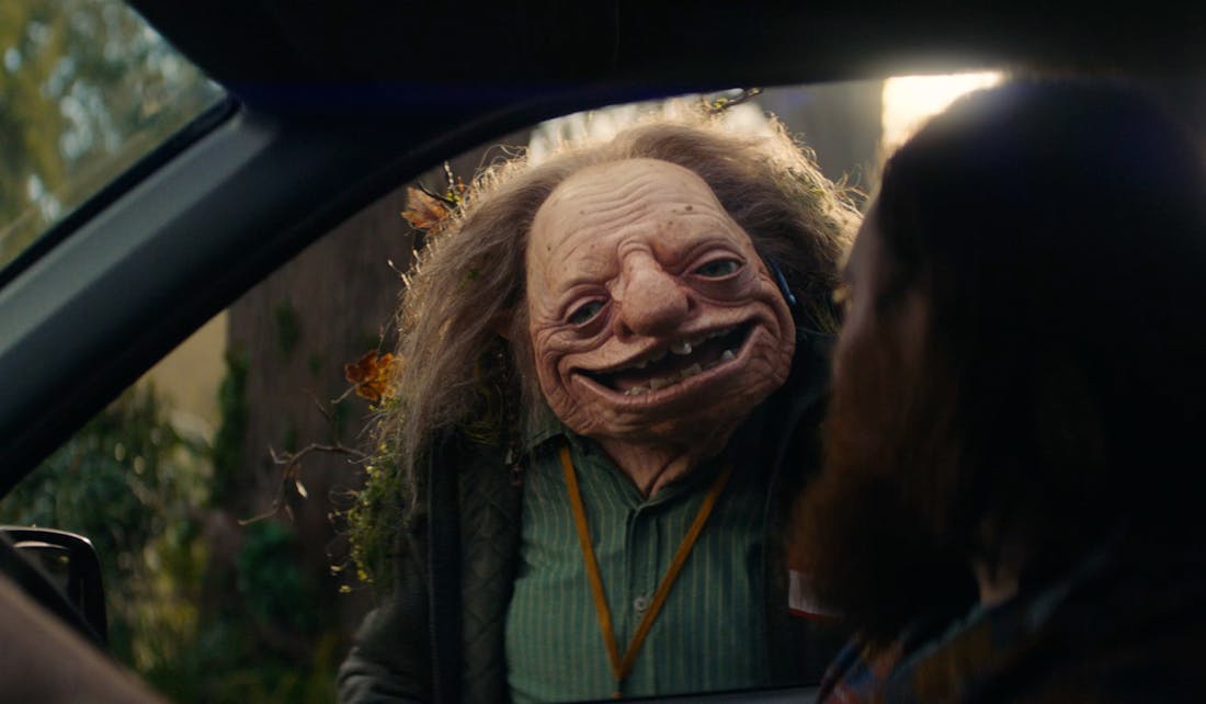 An image of a troll smiling, looking through a car window at the driver. 