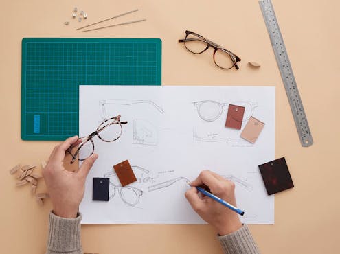 A hand holds a pair of Bailey Nelson glasses while sketching glasses with the other hand