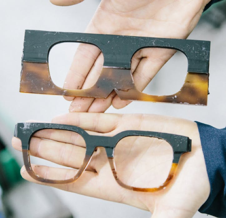 A hand holding a raw piece of acetate with lens holes next to a piece of acetate shaped into the front of a glasses frame for comparison