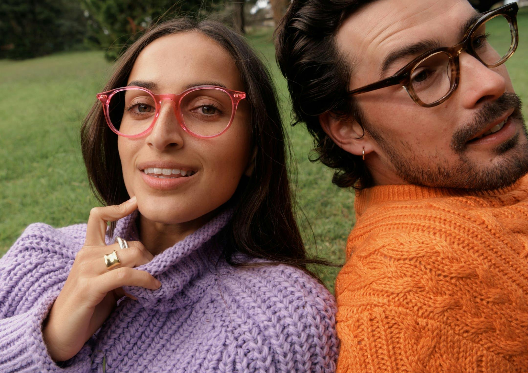 One person wearing hot pink round acetate Bailey Nelson Markova glasses in Hyper Pink and another in brown square acetate Bailey Nelson Harvey glasses in Desert Storm, in a field of grass.