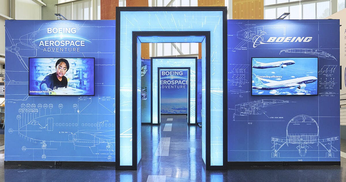 The entrance to the Boeing Aerospace Exhibit — a large, illuminated doorway with light blue walls on either side, decorated with monitor displays and blueprint-like sketches.
