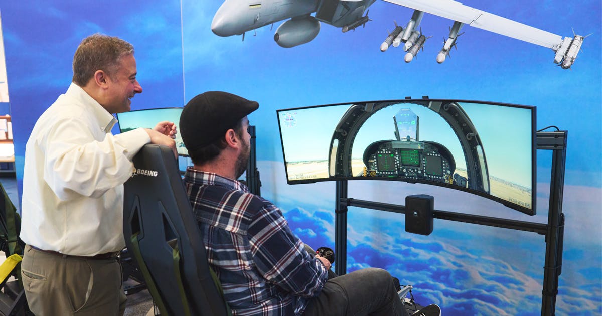 A man operates a flight simulator with dual curved screens showing a jet cockpit, while another person observes with a smile. A jet image decorates the wall.