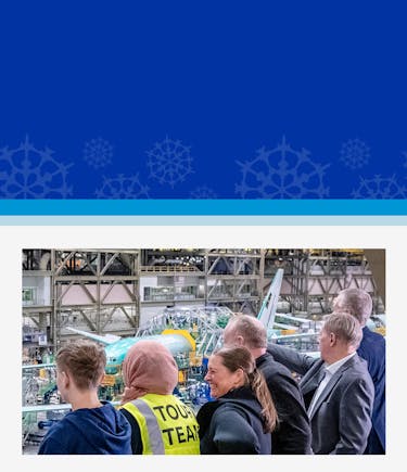 A group of people observes an airplane assembly inside a large hangar. A person in a yellow "Tour Team" vest is visible. The mood is curious and attentive.
