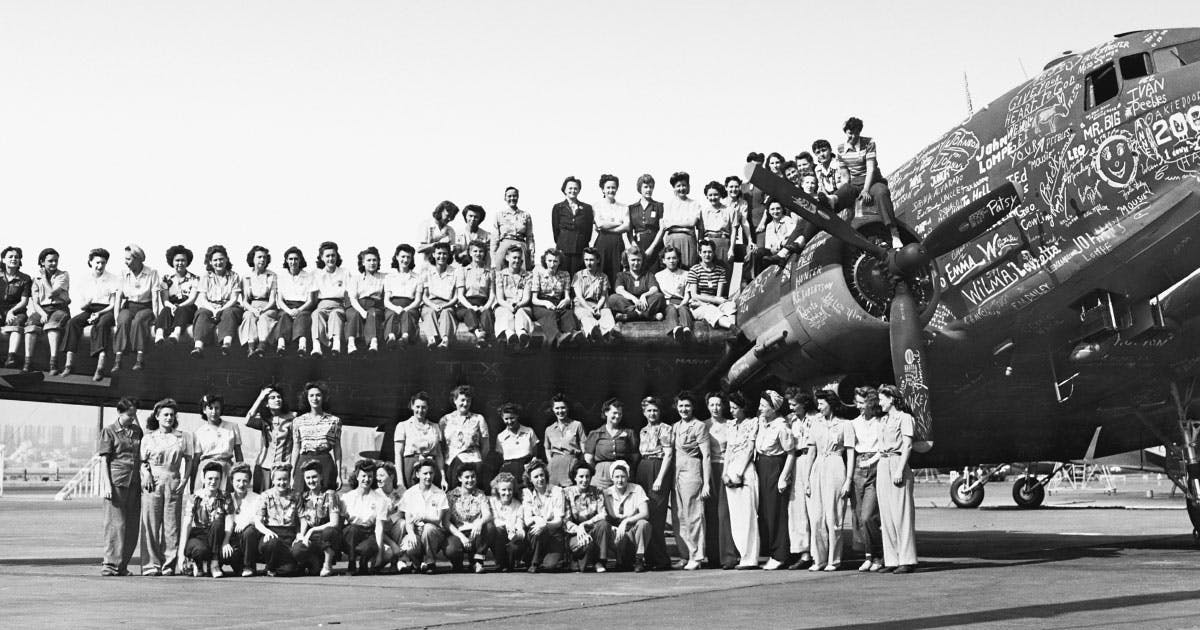 A black an white image of over 50 of our "Rosies" posed beneath and on top of the wing and fuselage of a Boeing war plane. 