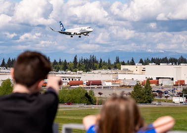 Kids observe a plane landing in Paine Field, with the Boeing Everett Factory and North Cascade mountains in the background.