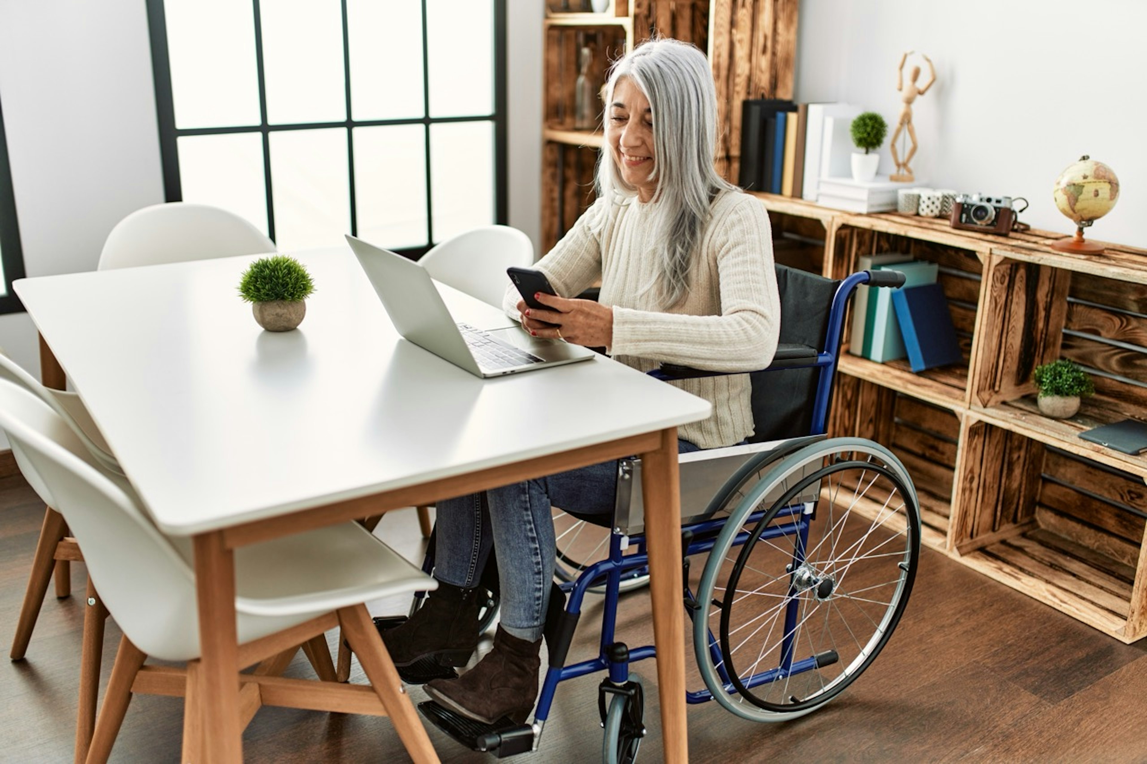 Person using a wheelchair sits at a dining table with a laptop, smiling while checking a smartphone in a bright home workspace.