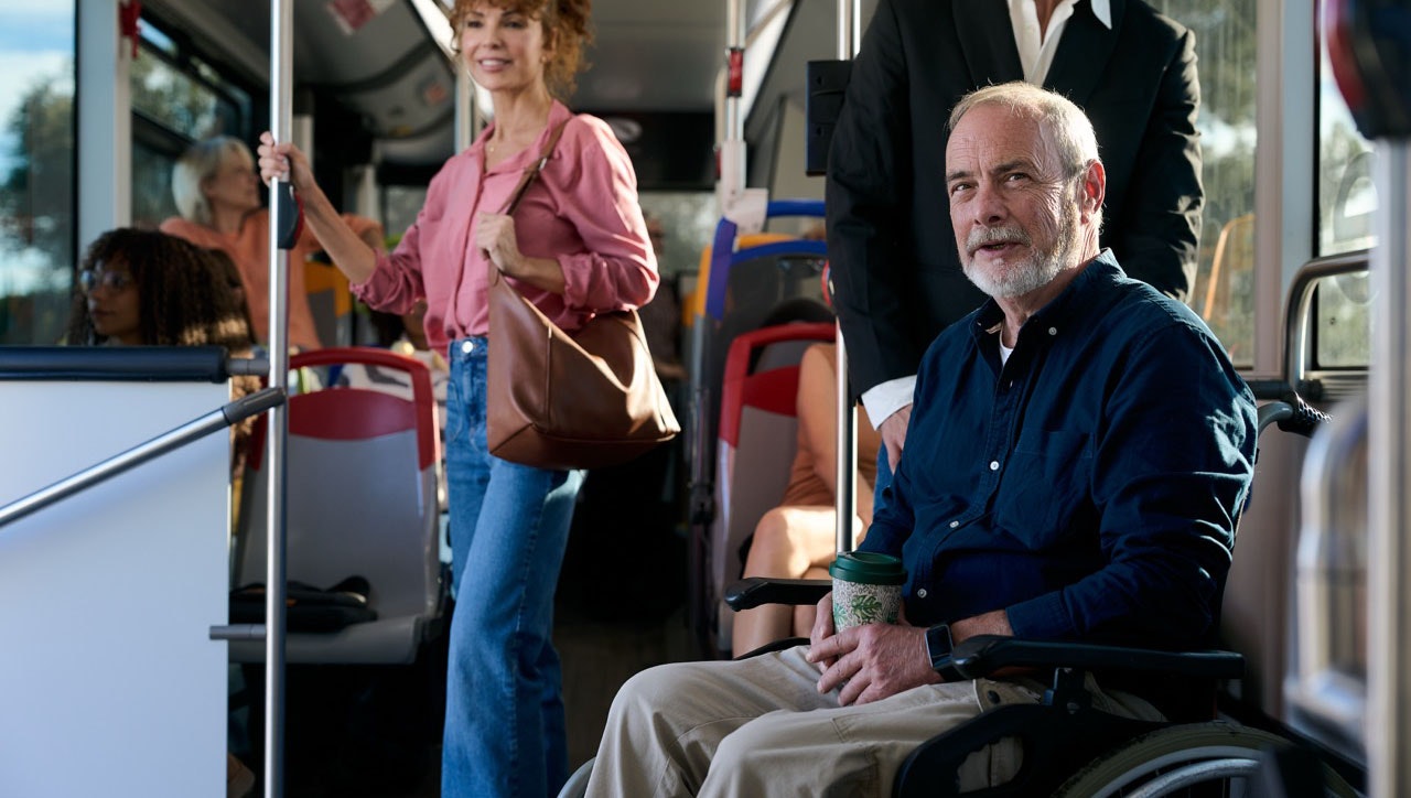 Passengers riding a city bus, including an older man using a wheelchair seated near the aisle, with other riders standing and seated around him.