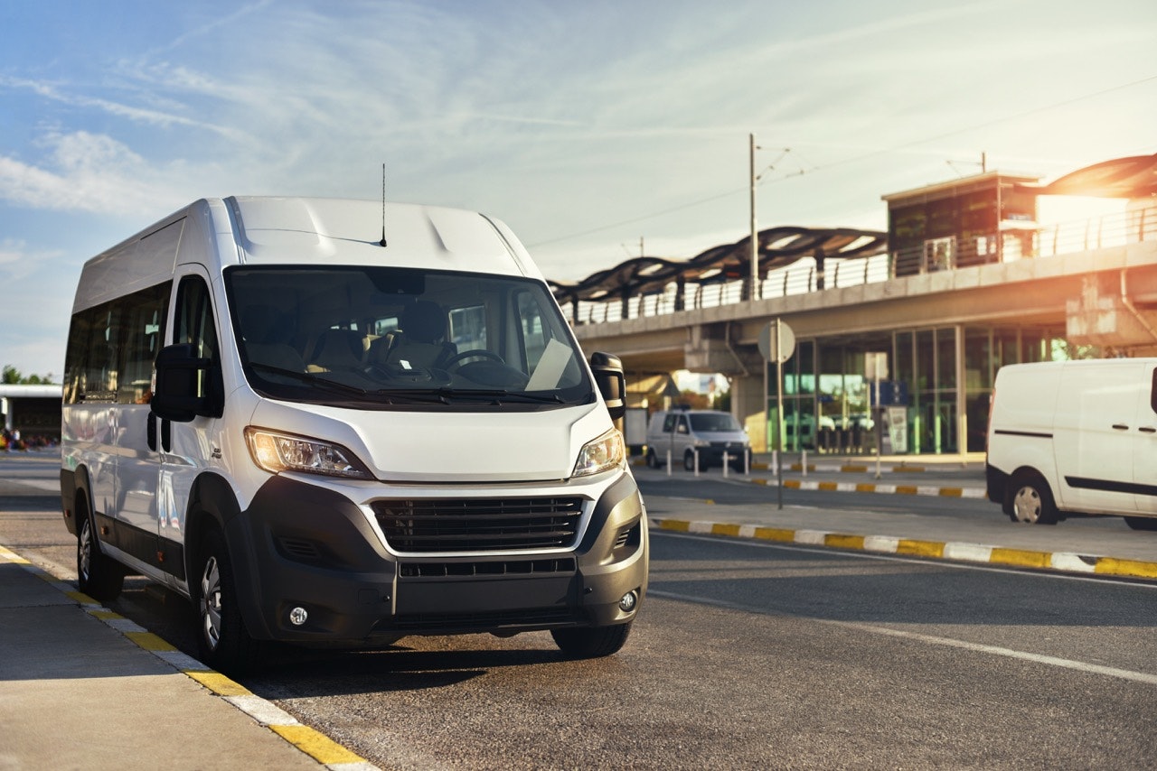 White shuttle van parked curbside near a transit station, positioned for passenger pickup during daylight.