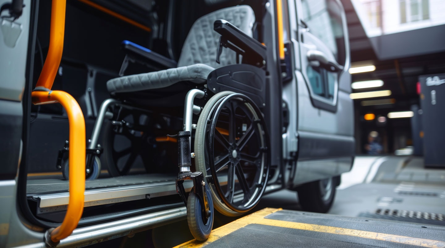 Empty wheelchair positioned on a deployed ramp at the entrance of an accessible transit van, showing interior handrails and securement area.