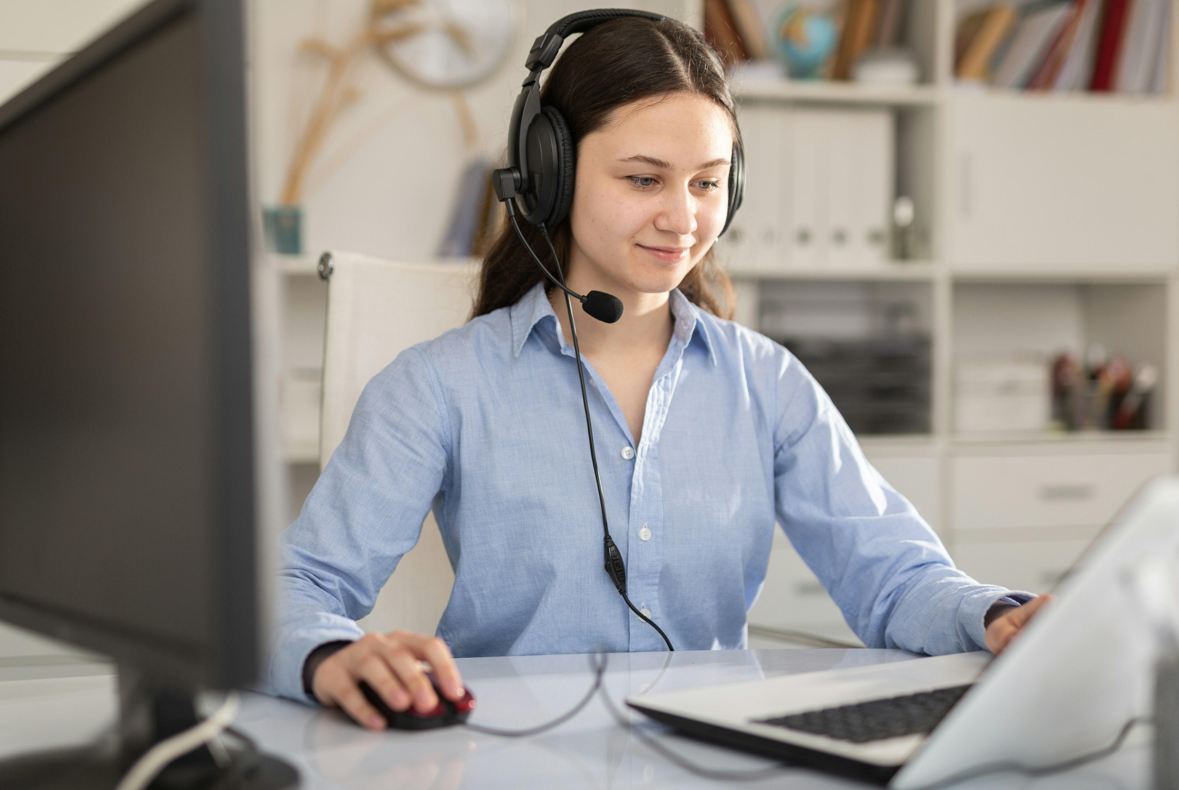 Customer support agent wearing a headset works at a desk, using a computer and mouse in a bright office setting.