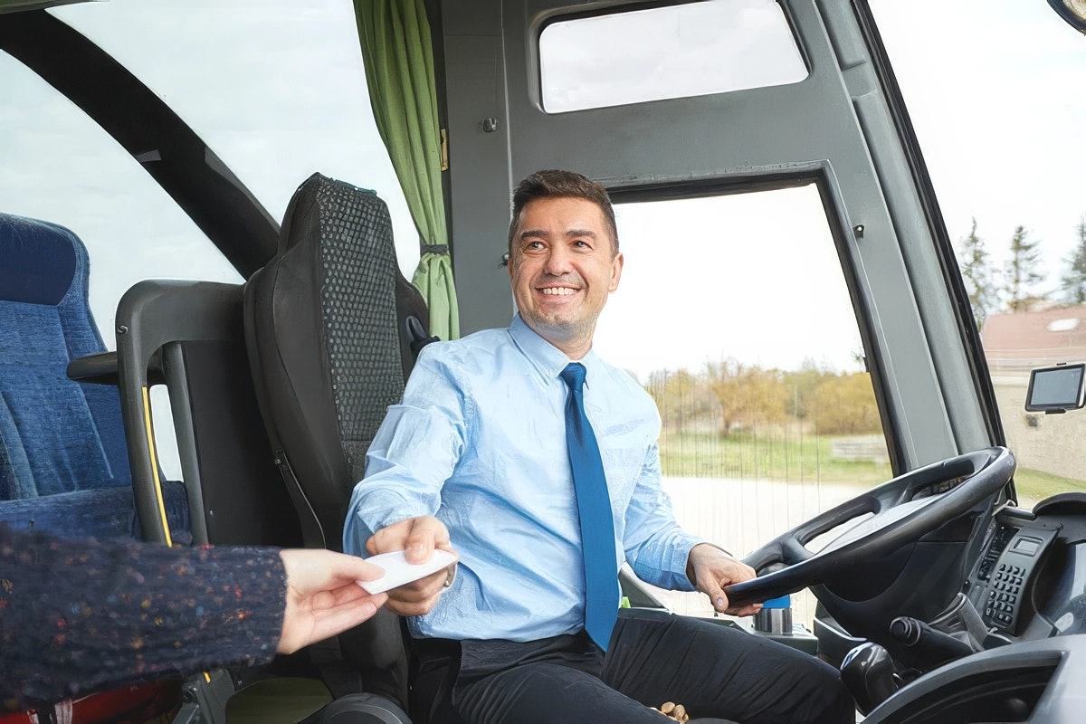 Shuttle bus driver smiles while accepting a fare card from a passenger at the front of the vehicle.