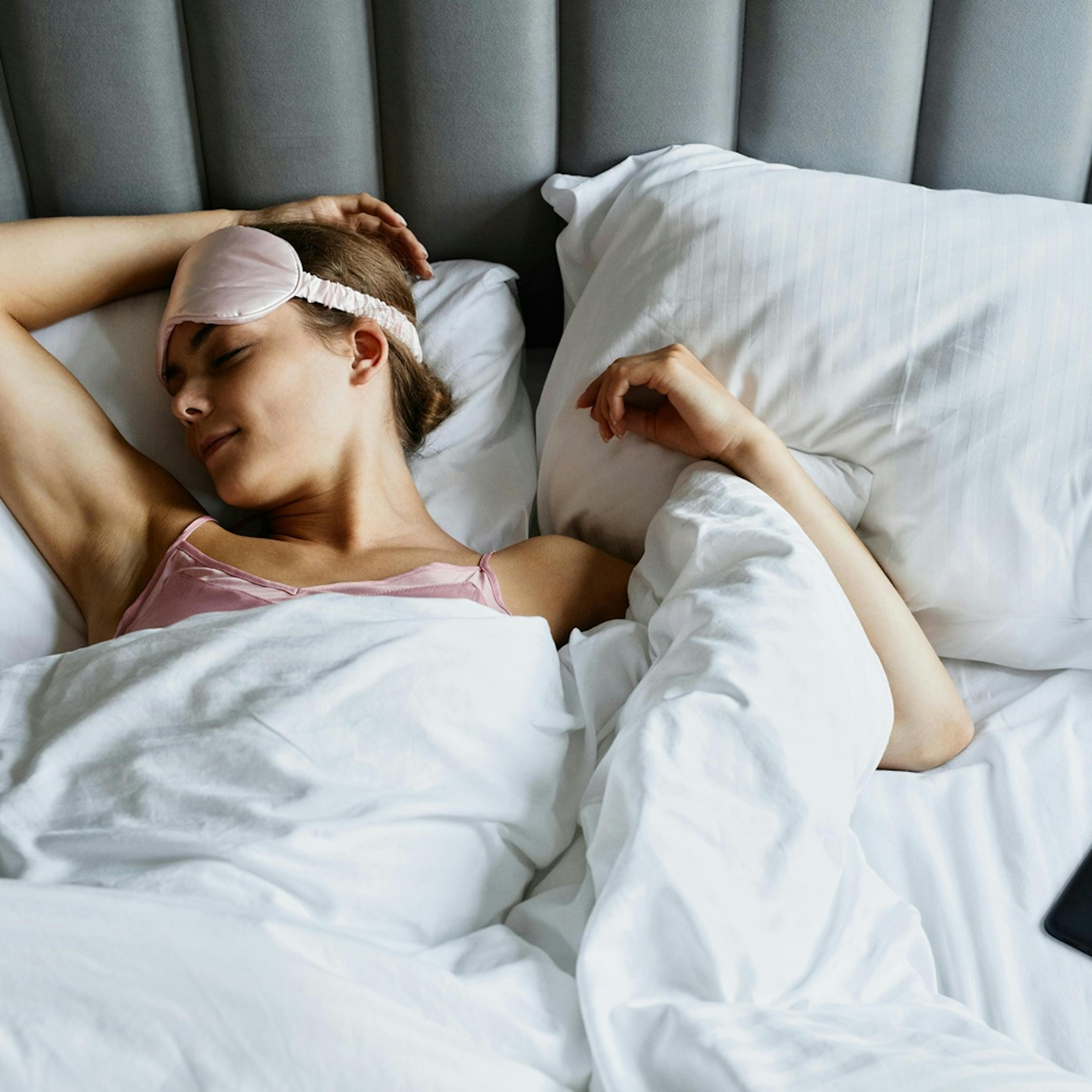 spící žena se škraboškou na čele, ležící v posteli s polštáři a peřinami, v dosahu mobilní telefon