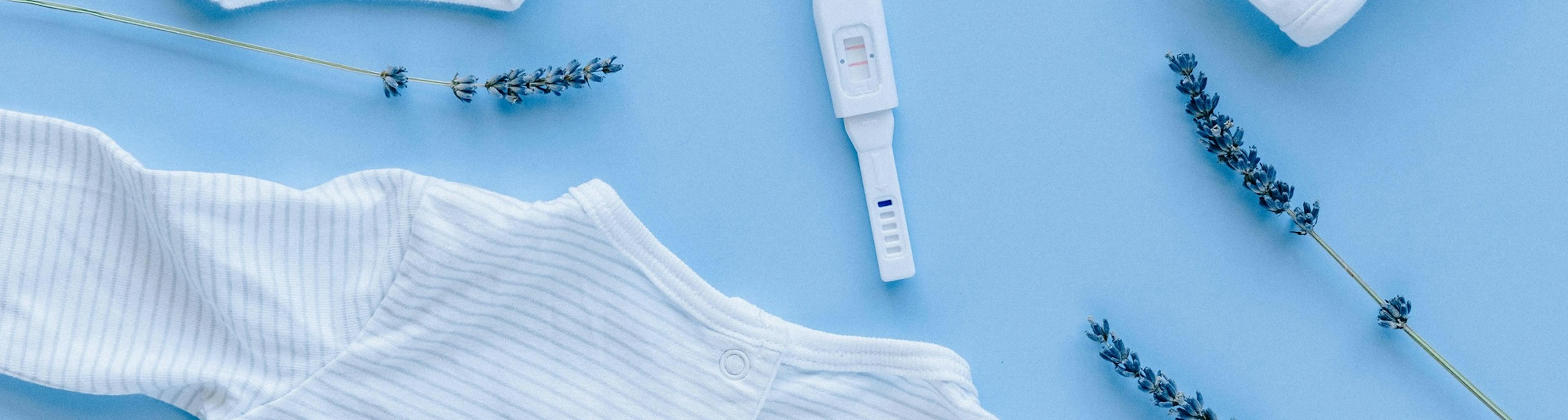 těhotenský test a okolo něj vyskládané oblečení pro miminko