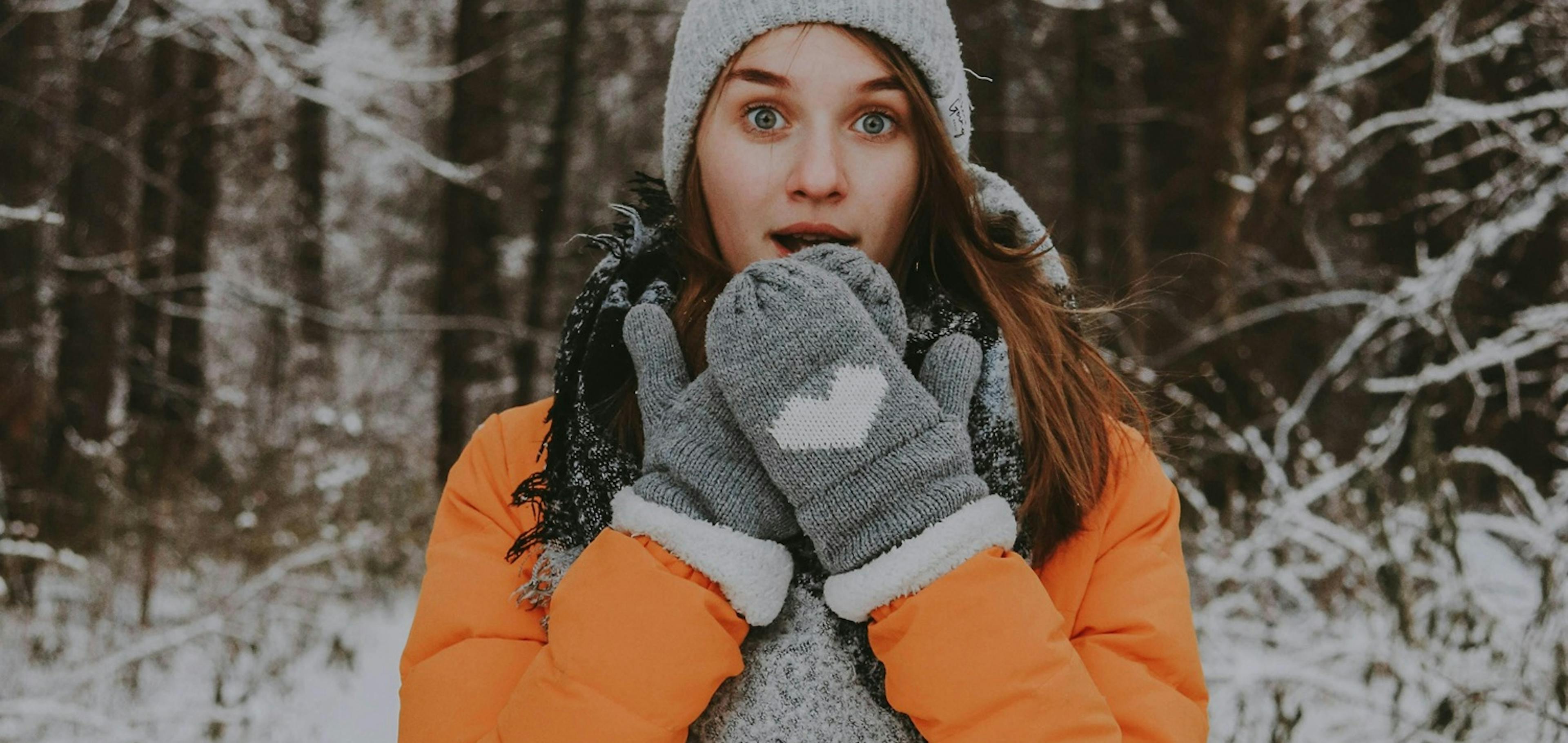 Zima, sníh, na pozadí zasněžený les, v popředí mladá ženy v zimním kabátu s čepicí a rukavicemi