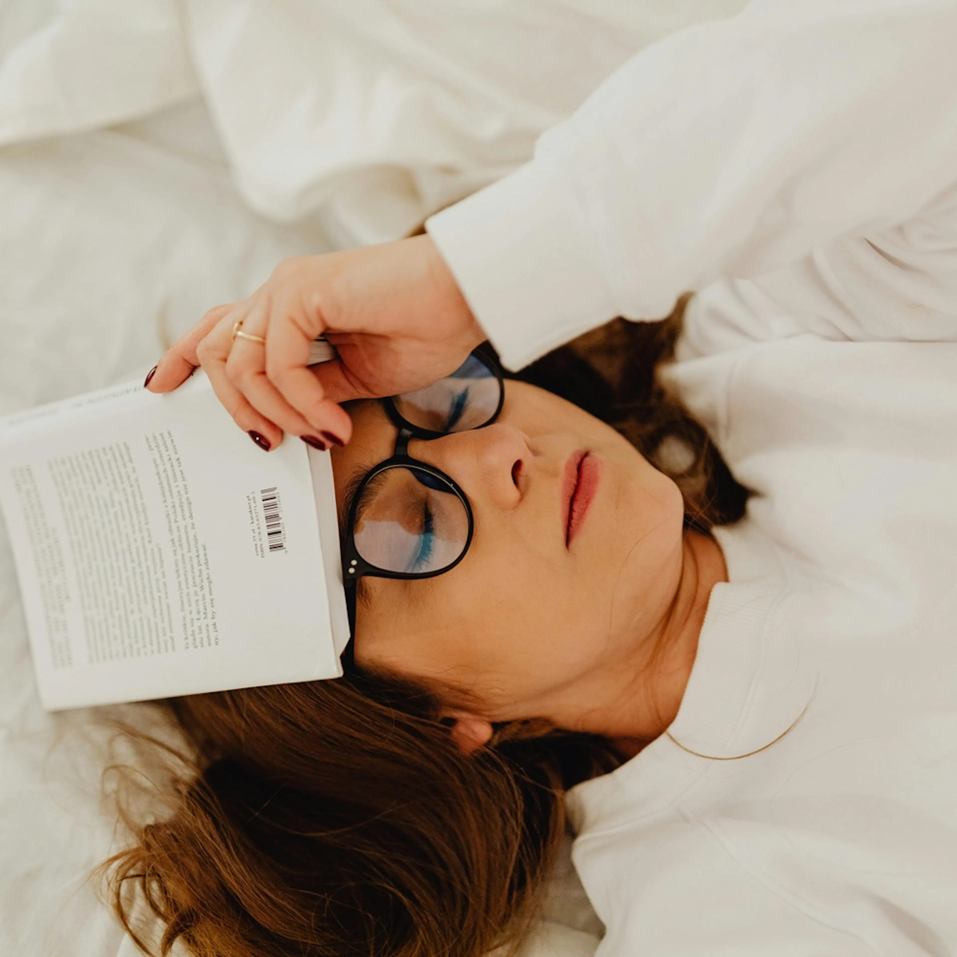 žena leží na posteli, má brýle a na hlavě knihu, má zavřené oči a je unavená