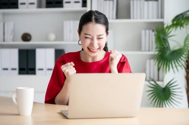 happy female professional in office background with pumped fists in front of laptop