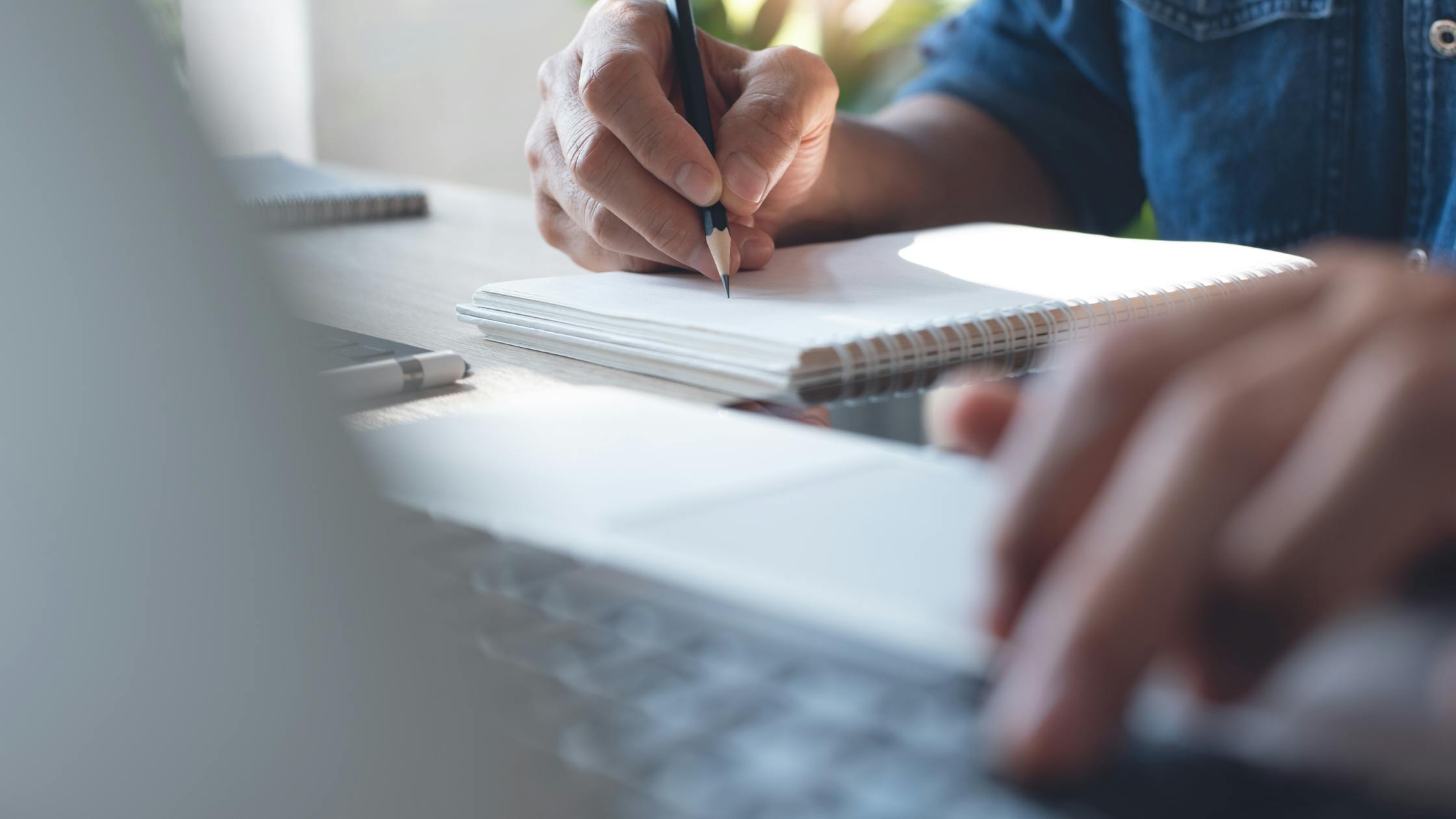 Individual writing in a book and working on a laptop