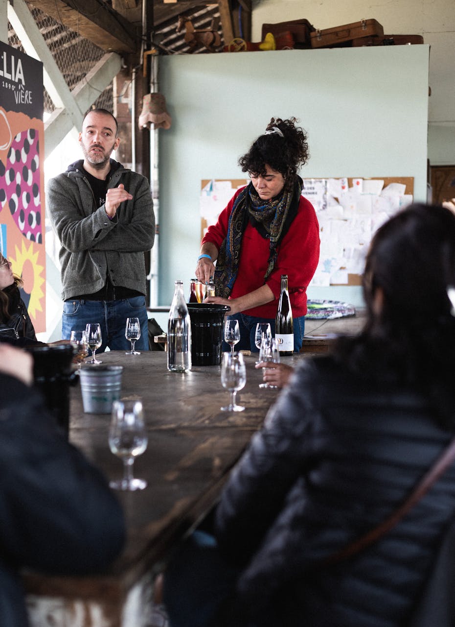 Céline Maguet qui débouche une bouteille derrière une table, avec des gens autours qui la regardent.