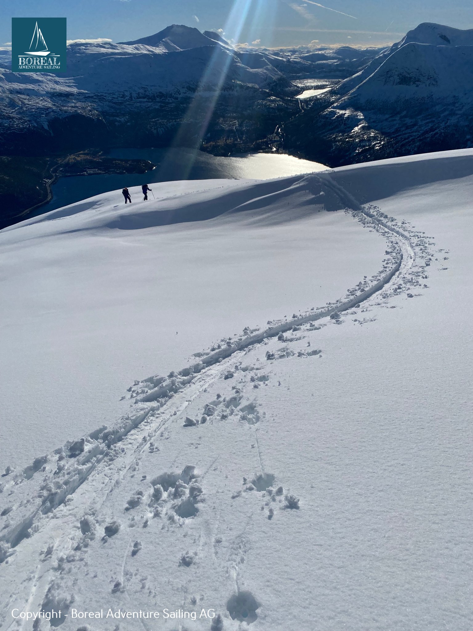 Ski & Sail, Bodø & Lofoten: winding skin track leading to two skiers above a shining fjord