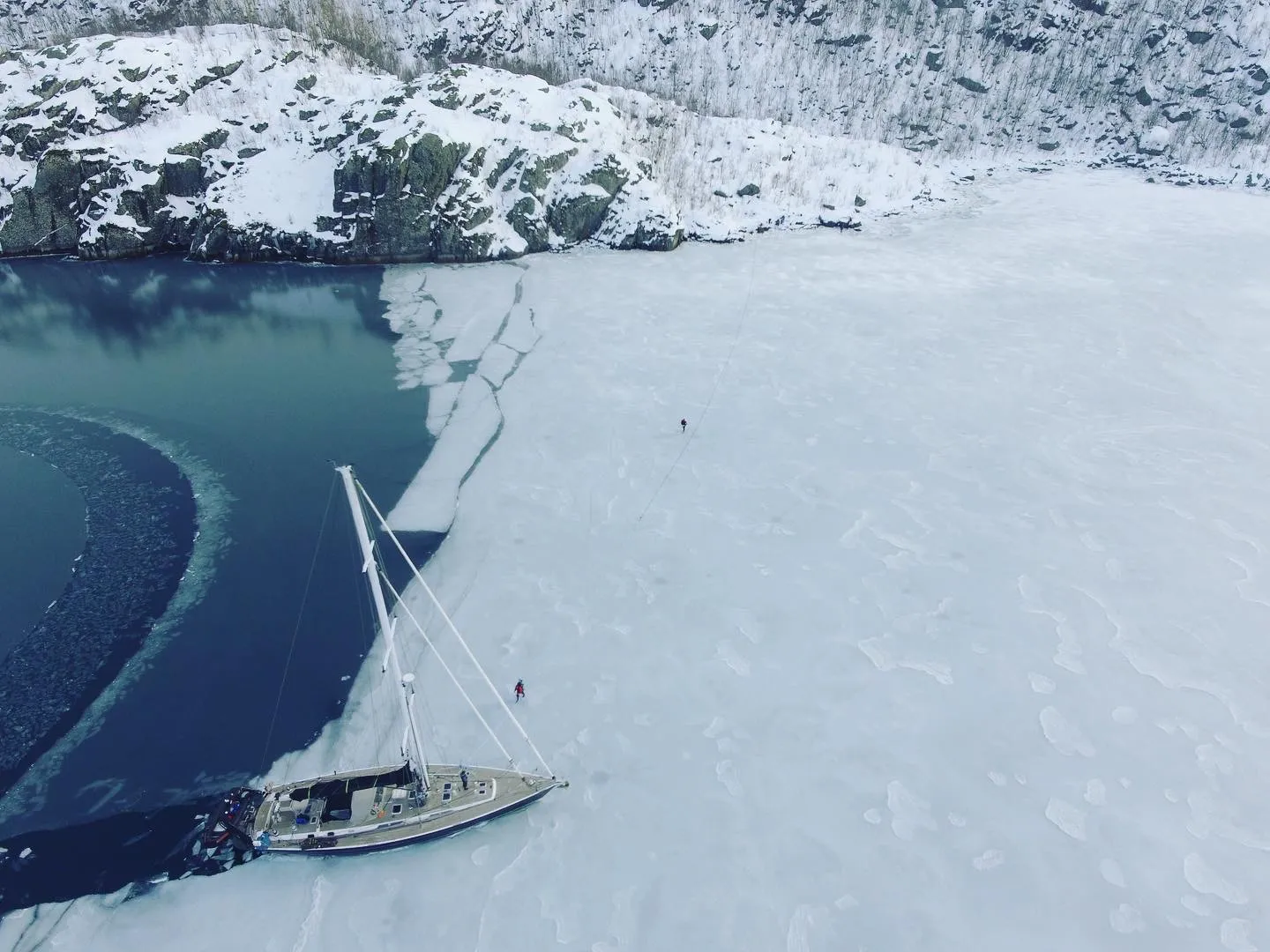 Sailing yacht nosed into fjord ice below snowy slopes, crew walking a mooring line ashore
