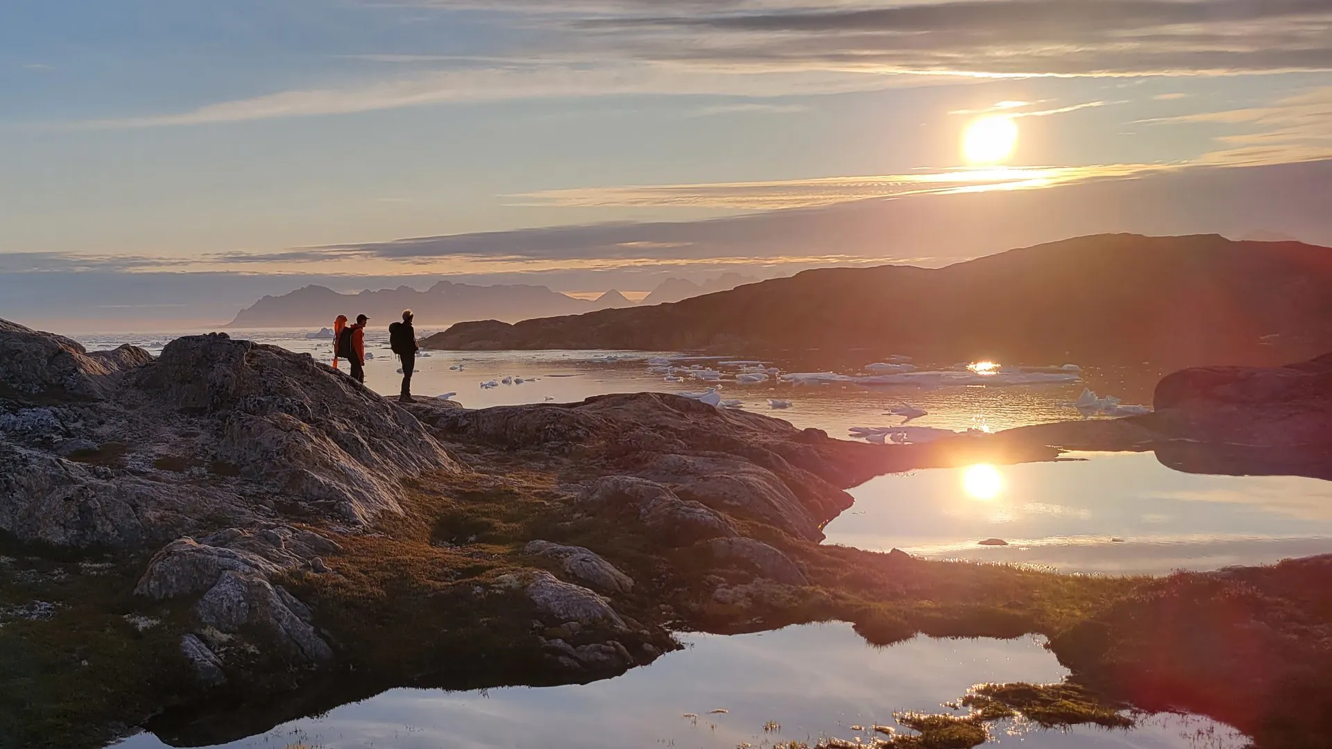 Voile & Exploration, Groenland de l’Est : randonneurs sur un rivage rocheux au coucher du soleil, le soleil se reflétant dans une mare de toundra