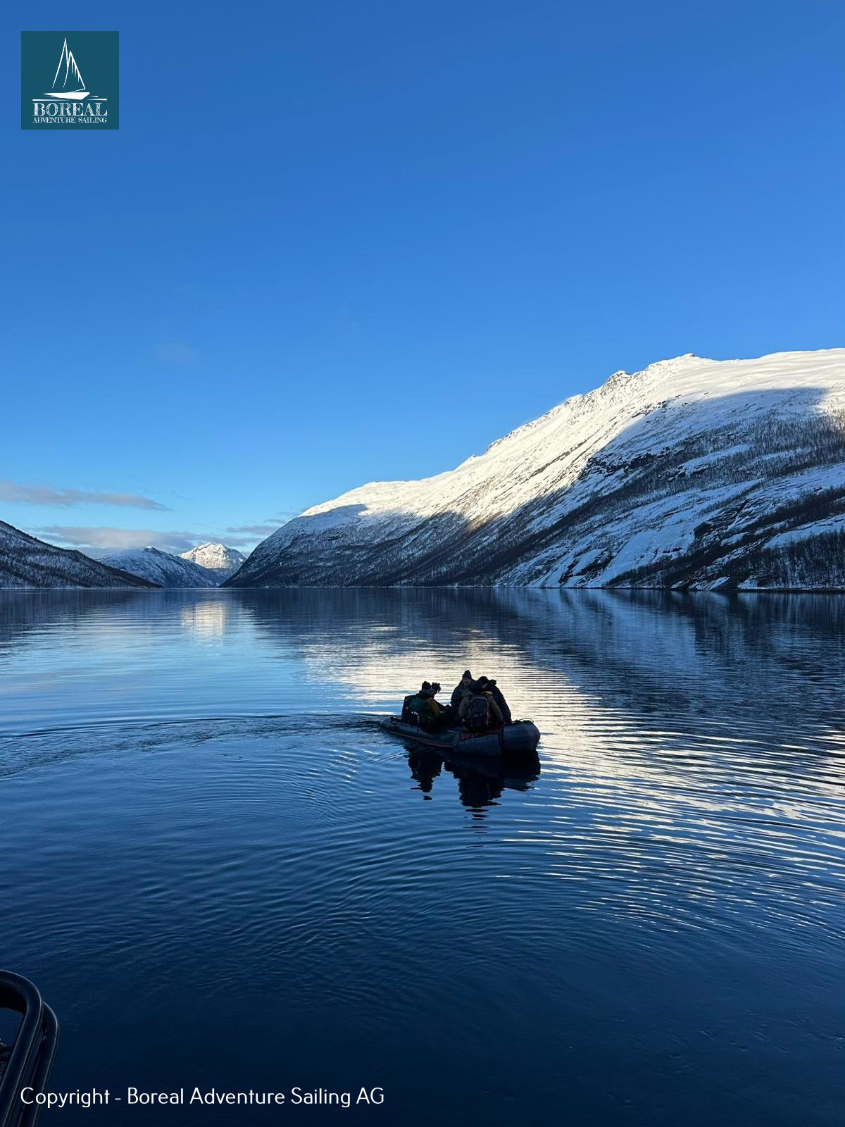 Ski & Sail, Bodø & Lofoten: dinghy crossing a mirror-calm fjord beneath a sunlit snowy mountain