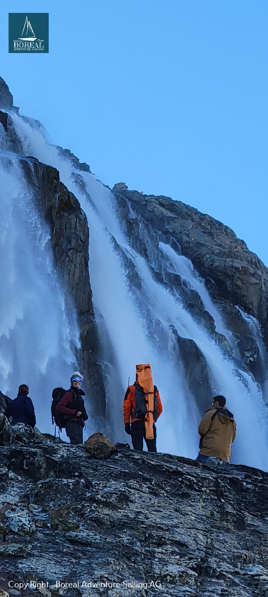 Sail & Explore, East Greenland: hikers pausing on rocks beneath a tall cascading waterfall