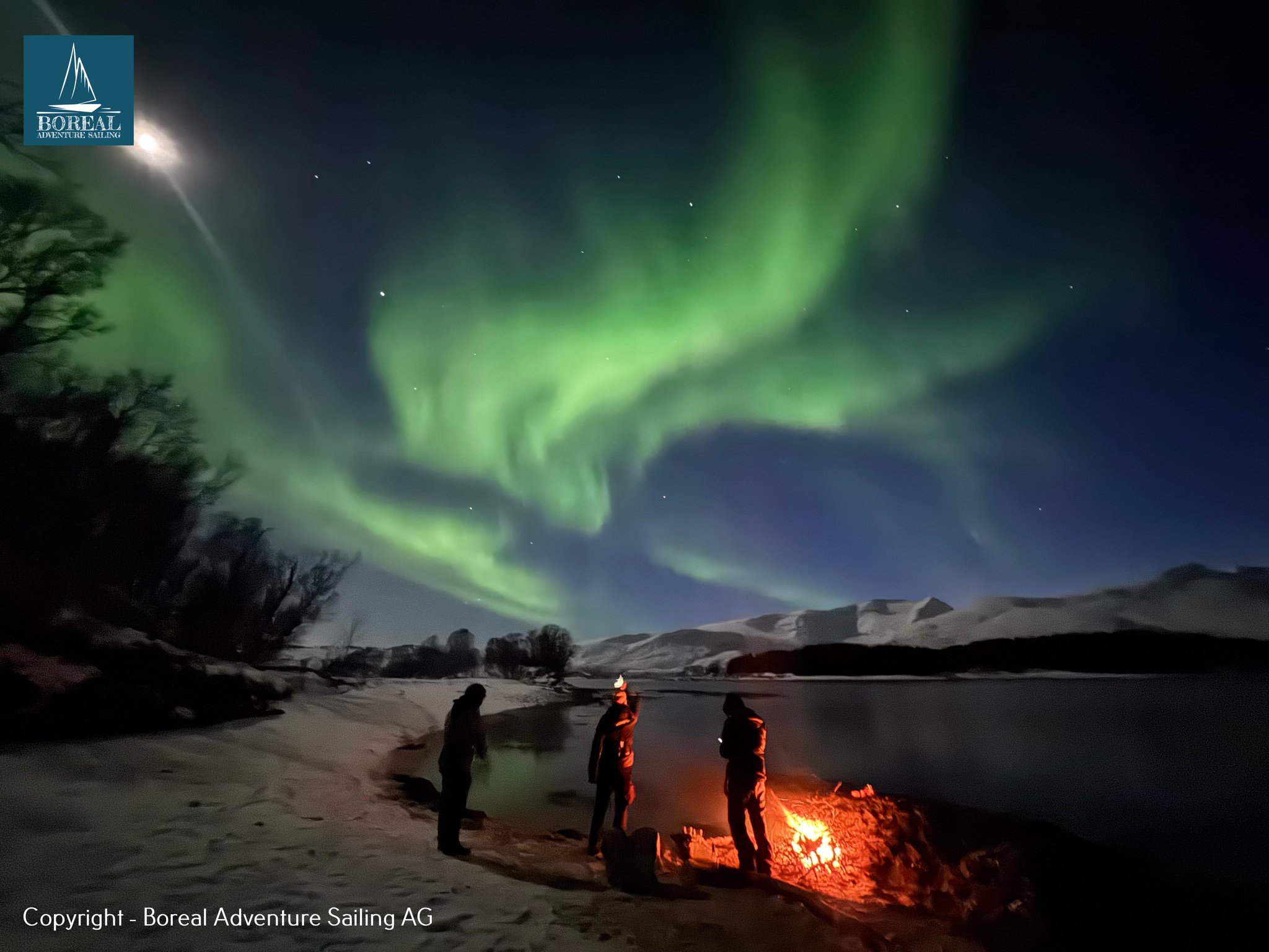 Ski & Sail, Lyngen Alps: northern lights above people around a beach campfire