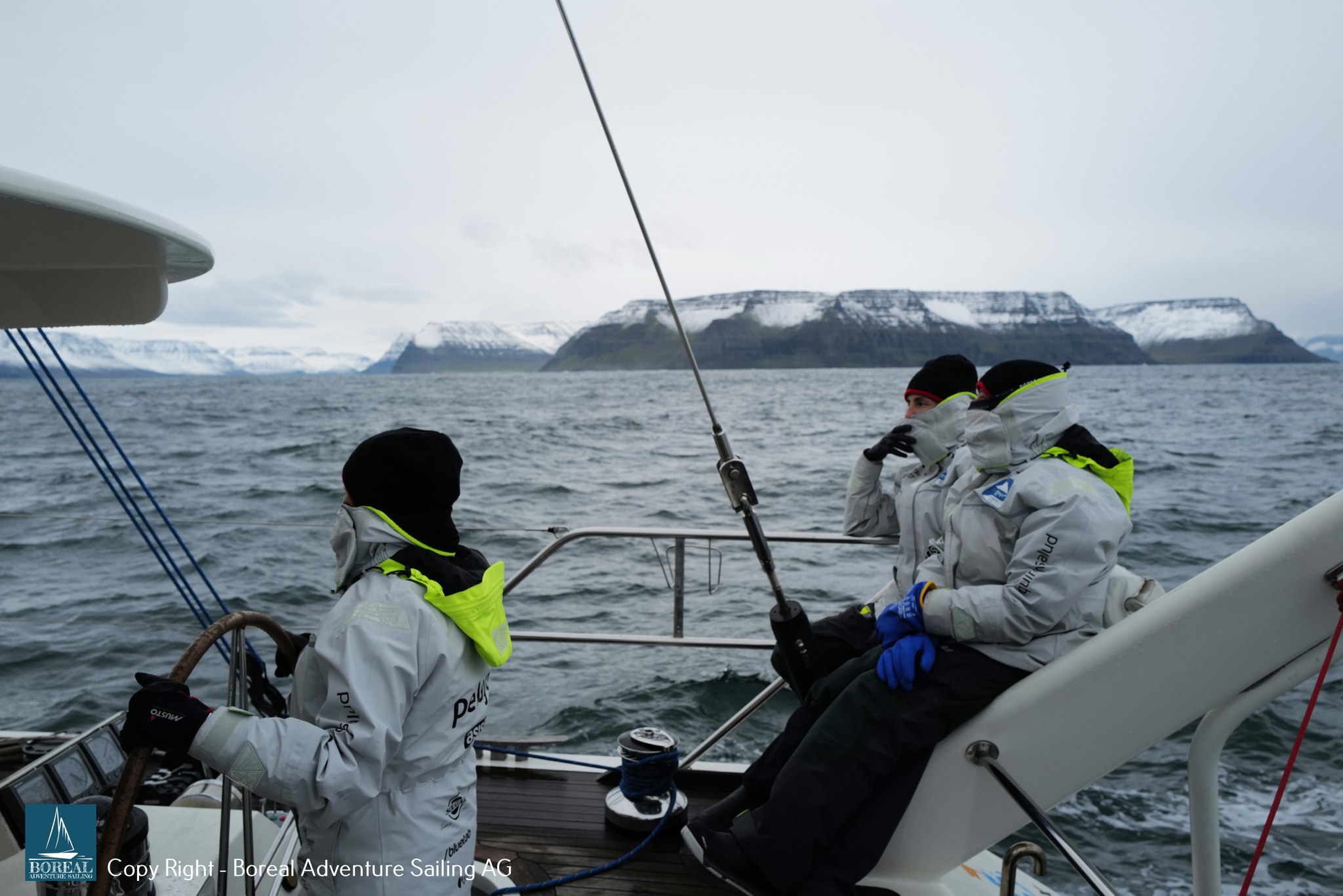 Offshore Sailing: crew at the helm with snow-streaked Icelandic mountains behind