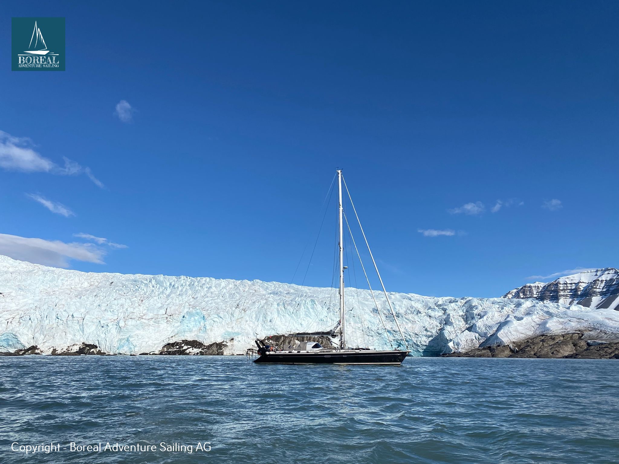 Sail & Explore, Svalbard: dark-hulled sailing yacht before a tidewater glacier under blue sky
