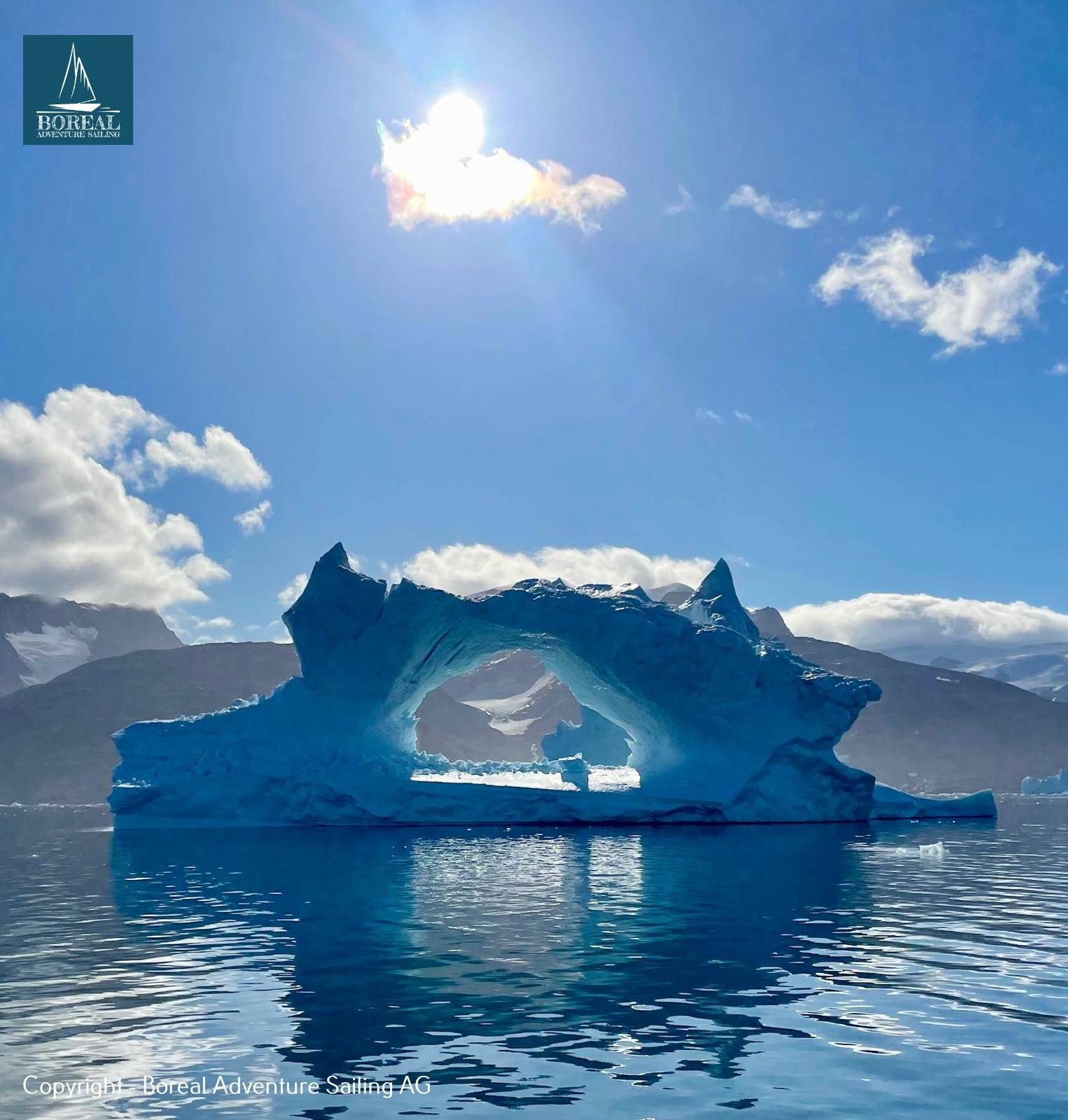 Sail & Explore, East Greenland: iceberg with a natural arch under sun and clouds