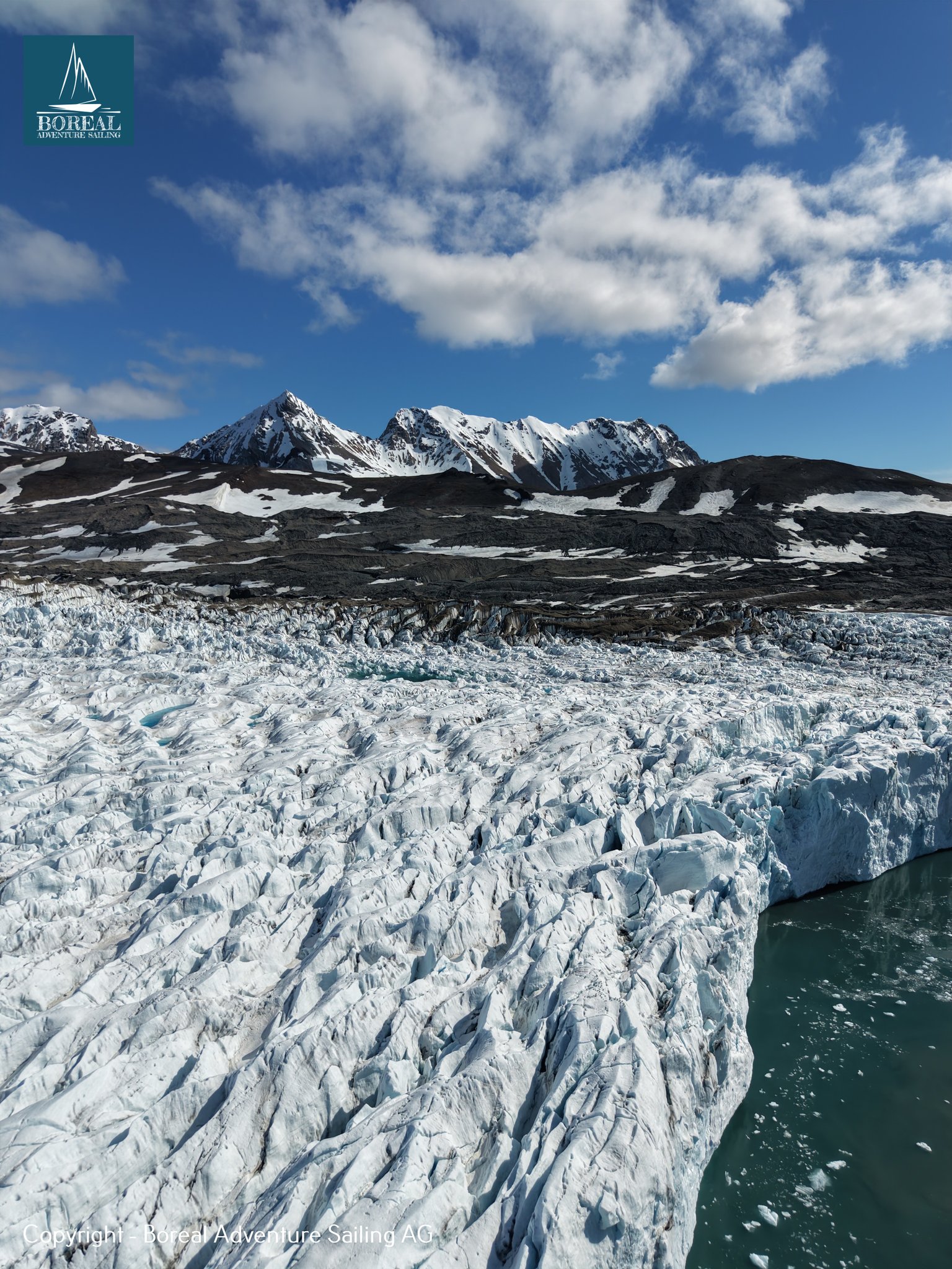 Sail & Explore, Svalbard: aerial view of a crevassed glacier front and peaks under blue sky
