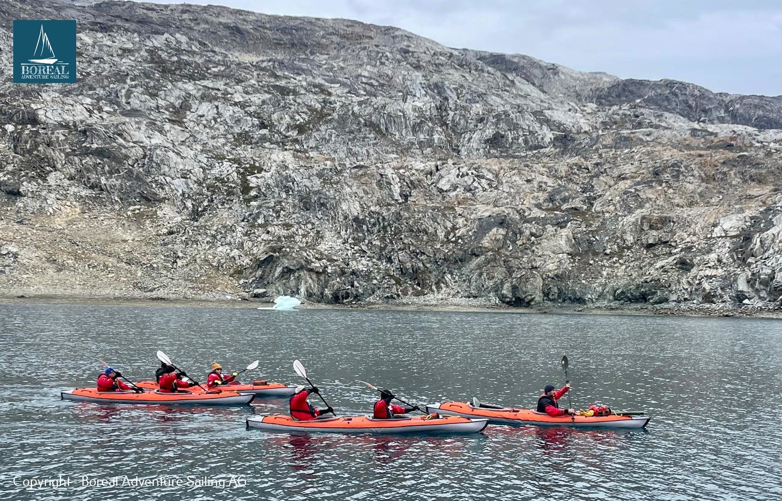 Sail & Kayak, East Greenland: group of red tandem kayaks paddling along a rocky shore