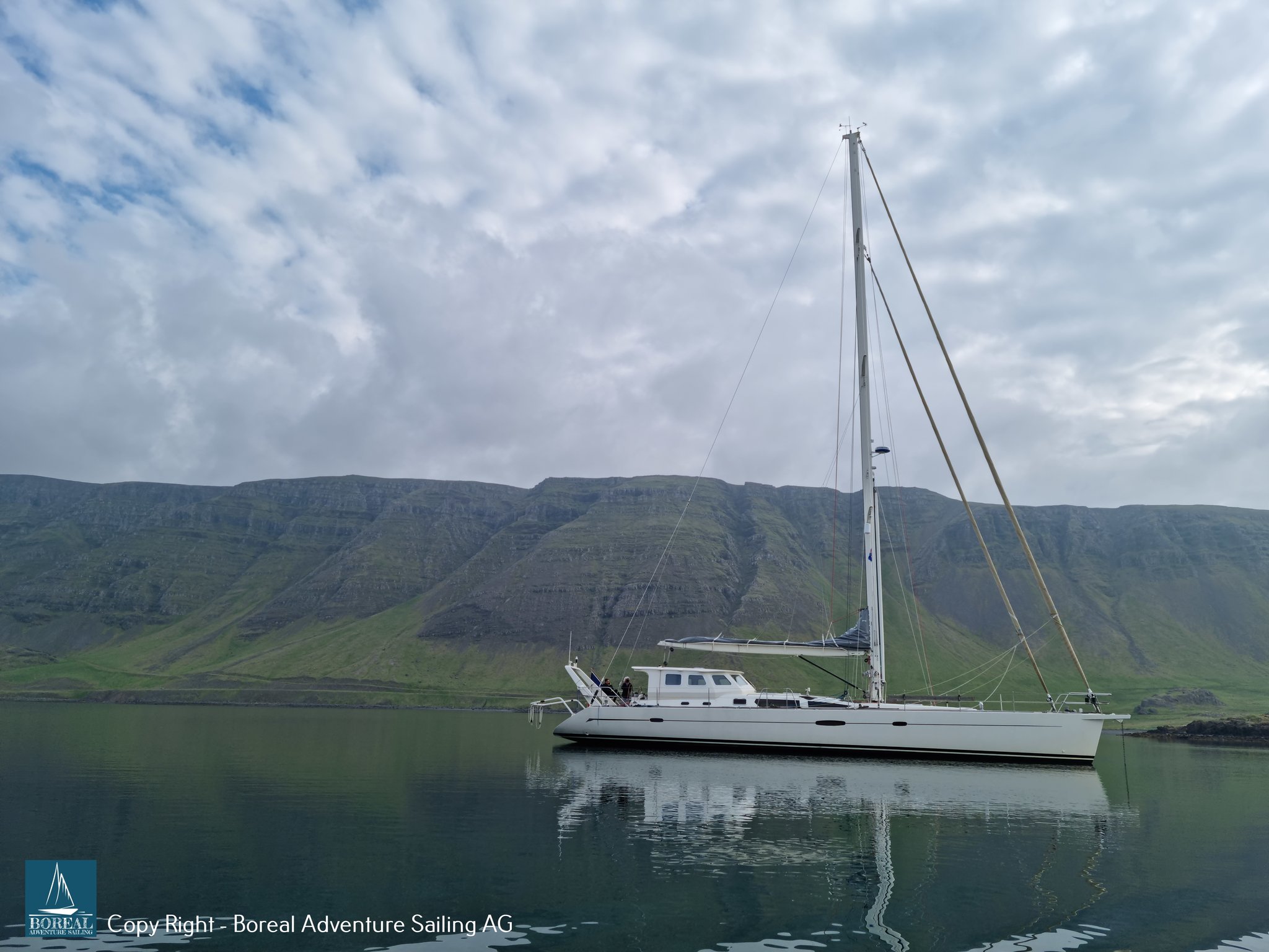 Offshore Sailing: sailing yacht at anchor in a mirror-calm fjord below green slopes