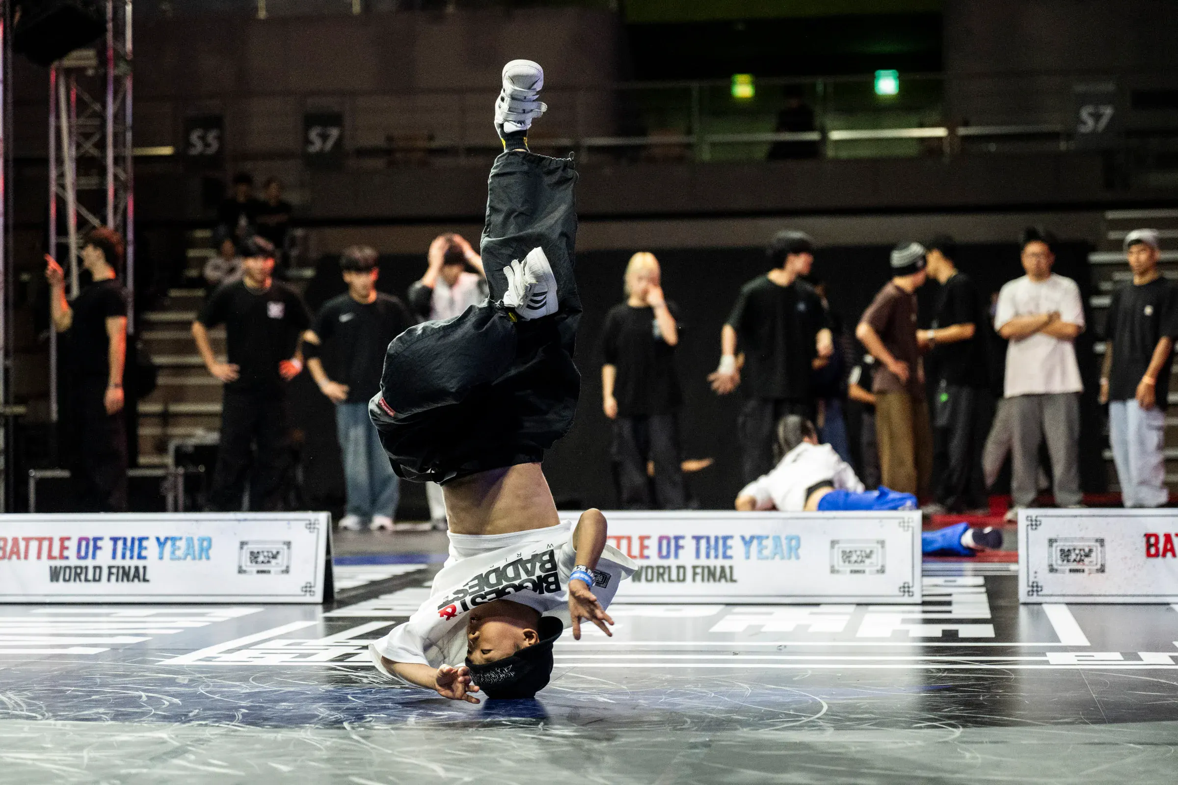 Freeze tête en bas pieds vers le ciel, t-shirt blanc 'BBOY BADDER', bandeau 'BATTLE OF THE YEAR' net derrière