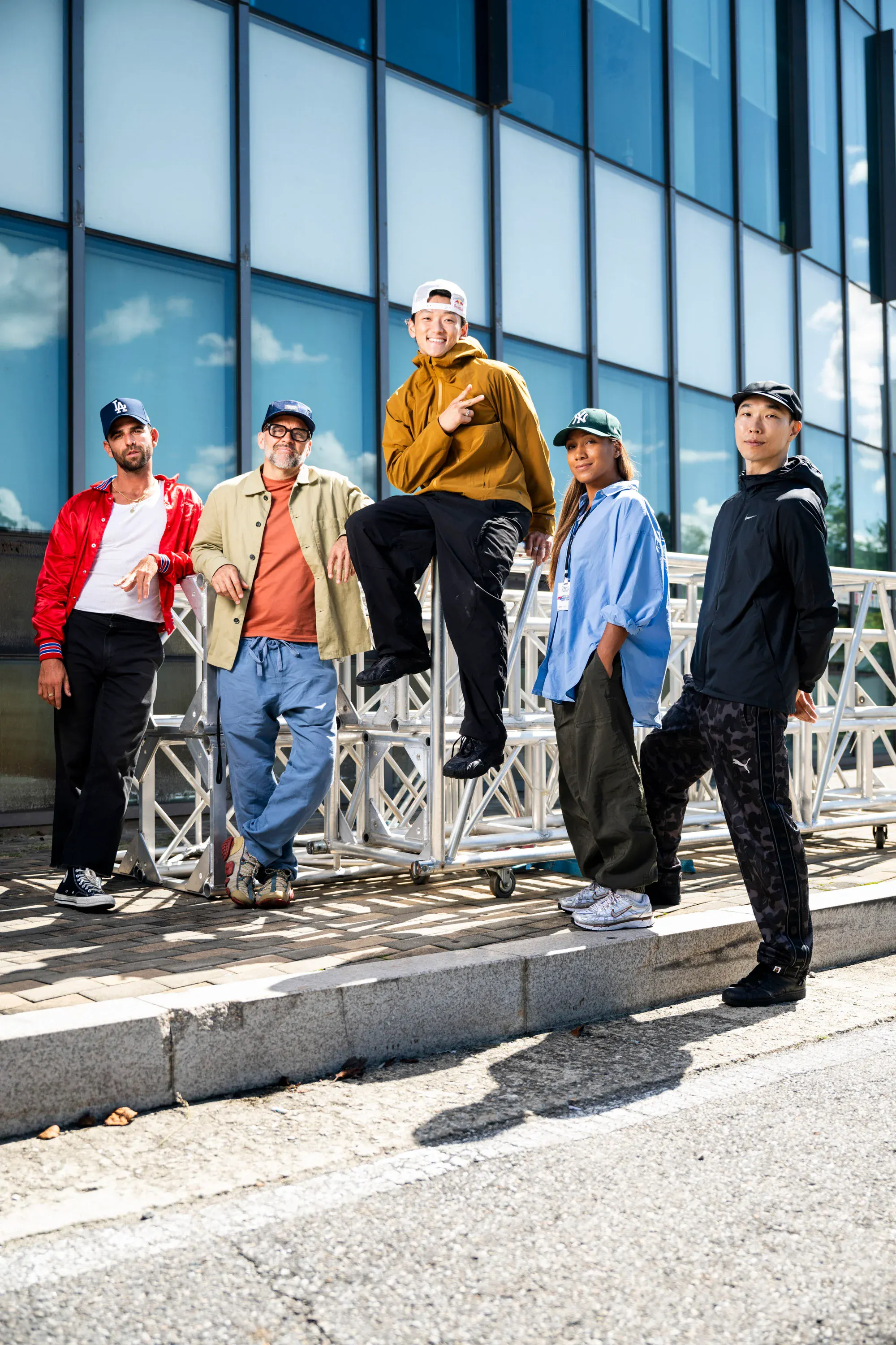Groupe judges/staff sur structure métallique, soleil, bboy en sweat moutarde au centre, vibe équipe
