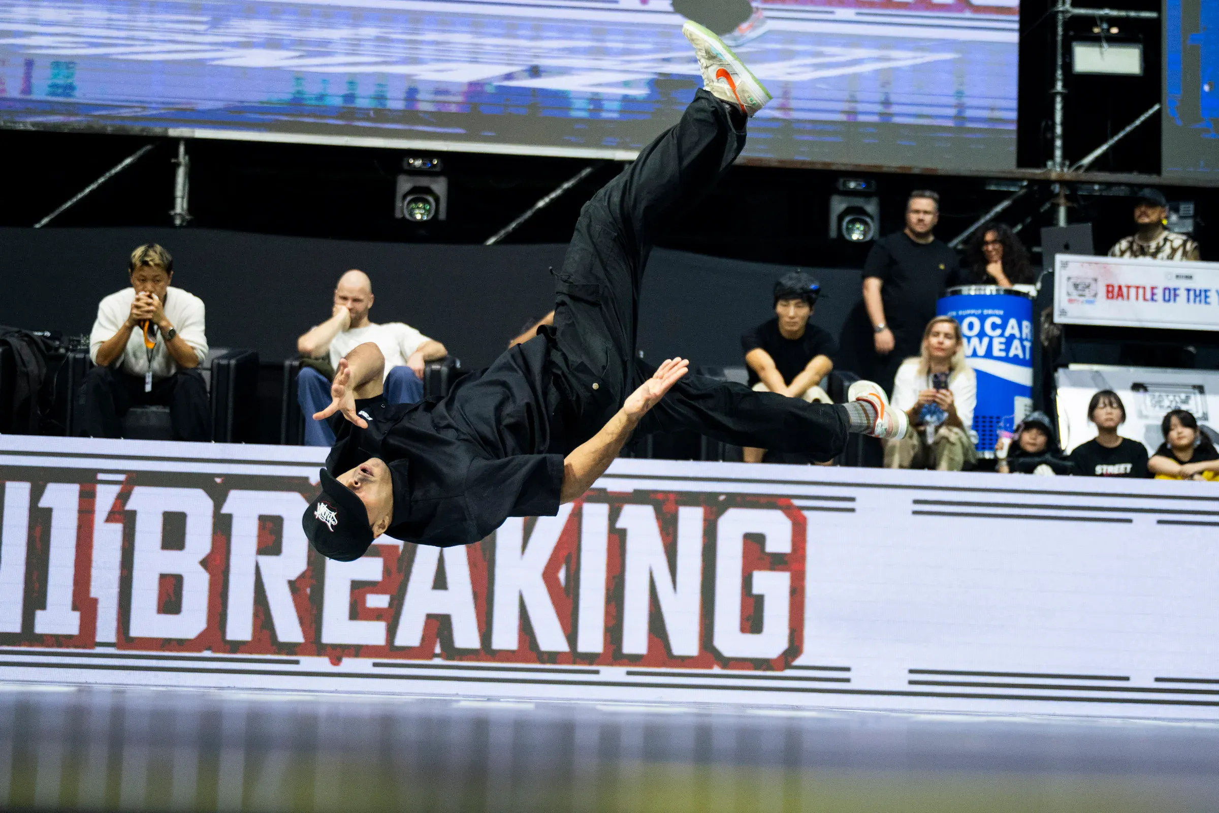 Bboy en saut horizontal complet bras en avant, écran 1ON1 BREAKING massif derrière, image puissante