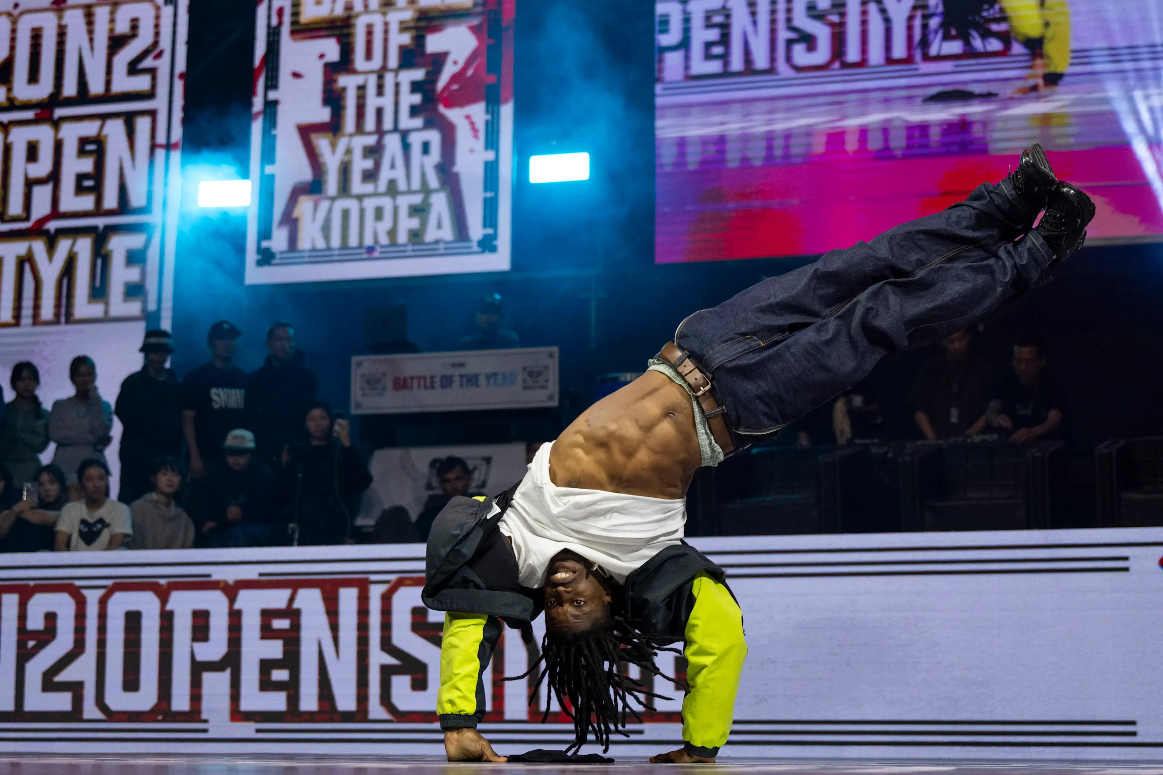 Bboy dreadlocks en handstand inversé jambes en l'air, abdos visibles, écran BOTY KOREA — image iconique