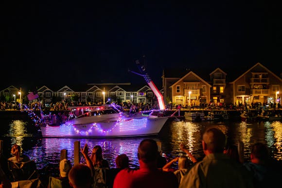 Crowd gathered along the riverbank watching a brightly decorated boat with lights pass by