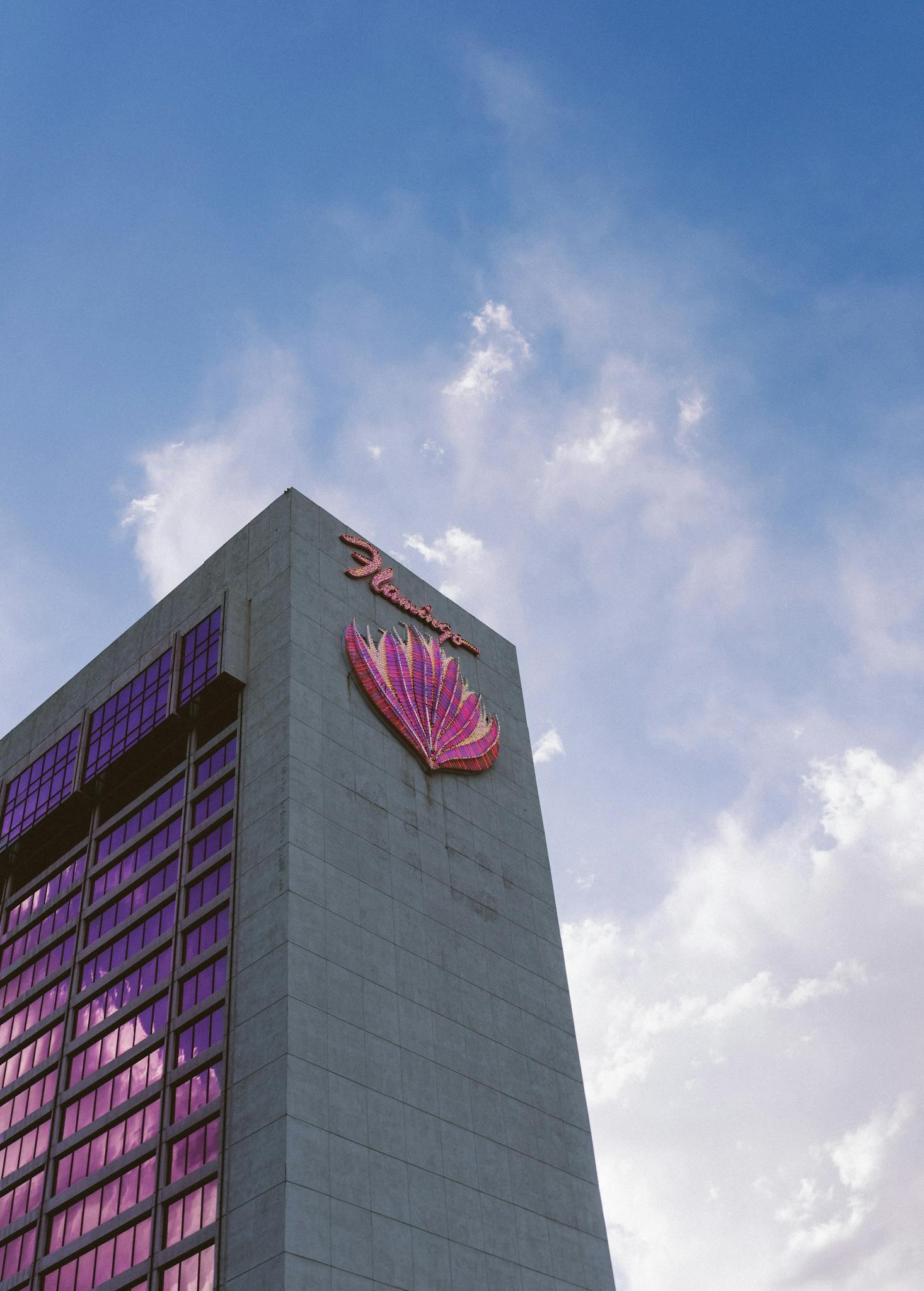 The grey tower of the Flamingo Las Vegas and the pink logo with blue sky and clouds behind it.