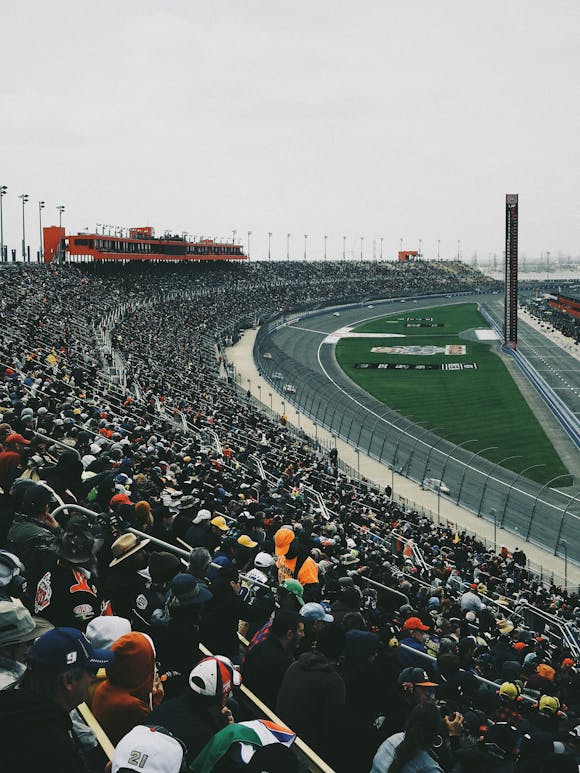 Huge crowd filling the stands at a car racing track, cheering as cars speed by