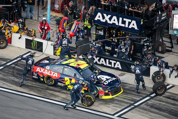 NASCAR pit crew services No. 24 Axalta car during a race, swiftly changing tires and refueling in a busy pit area