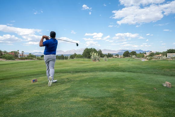 Golfer hitting a ball on a vast green golf field under a blue sky.