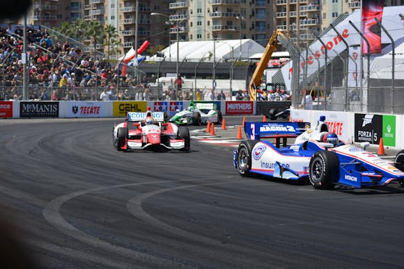 IndyCar vehicles speeding down a city track, surrounded by cheering fans in the stands 