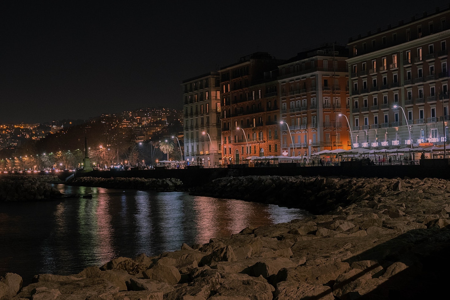 Lungomare Caracciolo waterfront and promenade with rocks by the water and lit-up buildings in the distance