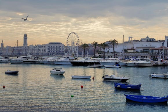 View of Bari's port with boats, Ferris wheel, palm trees, and the city skyline on a cloudy day